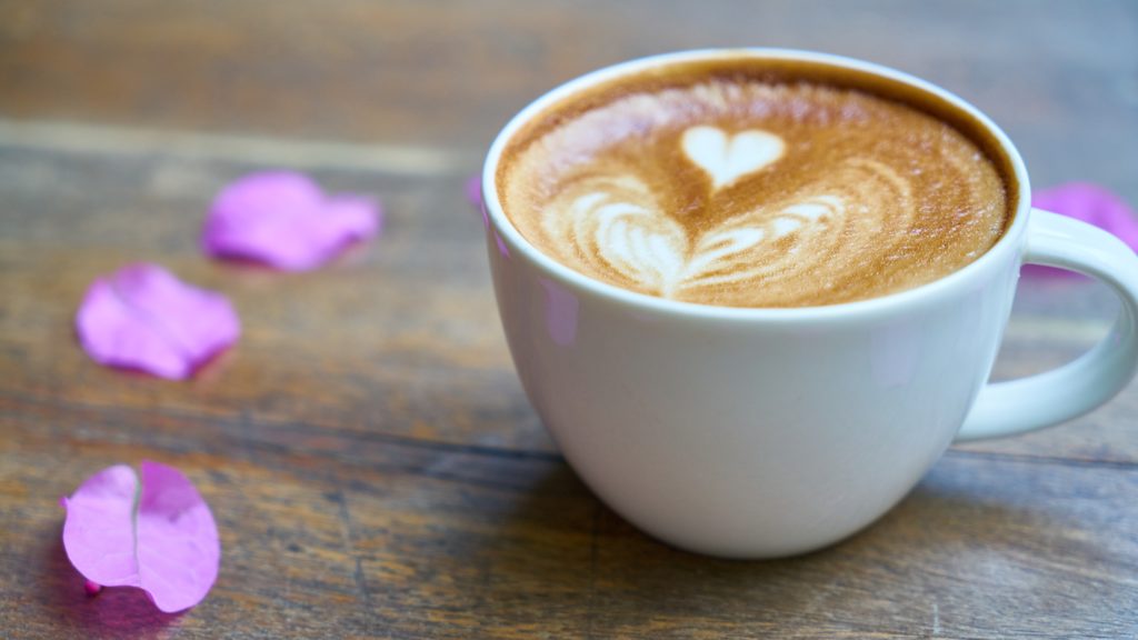 Wit kopje koffie met latte-art hartje, omringd door paarse bloemblaadjes op houten tafel